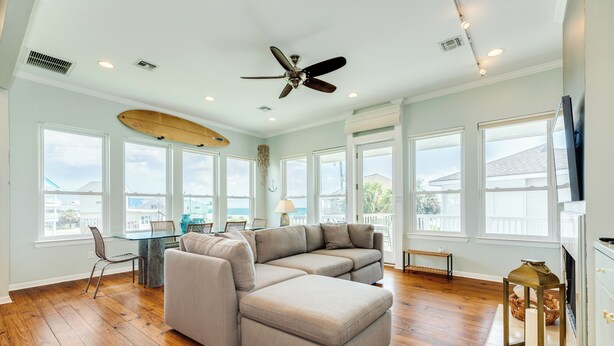 A spacious living room with a sectional sofa, wooden flooring, large windows, and a ceiling fan. - Bright & Airy 3rd Row Home w/ Gulf Views, 2 Decks, Outdoor Shower, Free Wifi (Galveston)