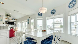Un comedor luminoso con mobiliario blanco, cojines azules, ventanas amplias y platos decorativos en las paredes.
