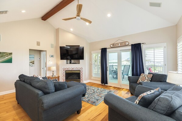 A living room with grey sofas, a flat-screen TV, a fireplace, and a sign reading 'BEACH HOUSE'.