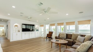 Ein geräumiges Wohnzimmer mit Holzboden, einem großen Fernseher, einem Deckenventilator und Meerblick durch große Fenster.