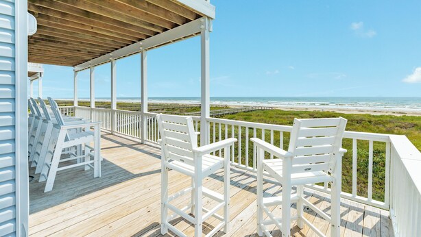 A wooden deck with white railings and chairs overlooking a beach.