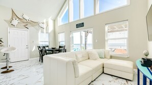 A spacious living area with a white leather sectional sofa, a dining table with chairs, and a bar area with a blue countertop. - Cozy Beachfront, Pet Friendly Home w/ Loft & Covered Patio - Steps from Beach! (Galveston)