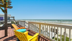 A wooden deck with yellow Adirondack chairs and a table, overlooking a beach with a white fence and a wooden pier. - Cozy Beachfront, Pet Friendly Home w/ Loft & Covered Patio - Steps from Beach! (Galveston)