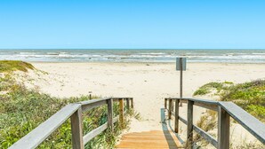 En træpromenade der fører til en sandstrand, med bølger i baggrunden.