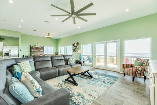 A spacious living room with a large gray sectional sofa, a wooden coffee table, and a colorful striped armchair. A ceiling fan and recessed lighting are visible. - Elevator, 2 Decks, Gulf Views all Bedrooms & More - Private Beachfront Home (Galveston)