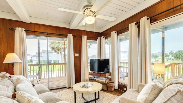 Una sala de estar acogedora con un sofá, una mesa de centro y un televisor. Hay cortinas, un ventilador de techo y vistas al exterior.