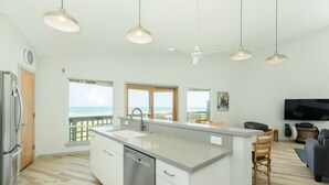 Una cocina moderna con una isla grande, armarios blancos y un lavavajillas integrado. Hay una zona de comedor con una mesa y sillas de madera, y un televisor de pantalla plana montado en la pared.