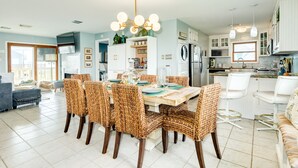 A spacious dining area with a large table and wicker chairs, a kitchen with white cabinets and a built-in oven, and a living area with a sofa and a TV. - Willy's Place: 3rd Row Gulf View Home with Updated Interior & Rooftop Sun Deck (Galveston)
