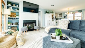 A living room with a fireplace, grey sofa, and a bookshelf. - Willy's Place: 3rd Row Gulf View Home with Updated Interior & Rooftop Sun Deck (Galveston)
