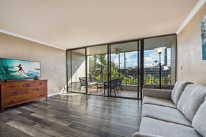 Un salon doté d’un plancher en bois, d’un canapé gris, d’un téléviseur posé sur un meuble en bois et d’une porte-fenêtre coulissante donnant sur un espace extérieur avec une table et des chaises.