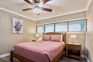 Une chambre à coucher comprenant un lit, deux tables de chevet, un ventilateur de plafond, une fenêtre avec des stores et un tableau accroché au mur.