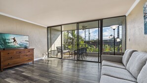 A living room with a wooden floor, a grey sofa, a TV on a wooden cabinet, and a sliding glass door leading to an outdoor area with a table and chairs. - Kalani Kai#104 1st Floor, Remodeled Unit in Heart of Kona Town! Best location (Kailua Kona)