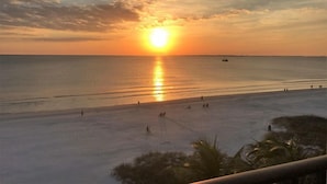 Ein Strandbild mit Sonnenuntergang über dem Meer, Menschen schlendern am Ufer entlang und ein Boot ist in der Ferne sichtbar.