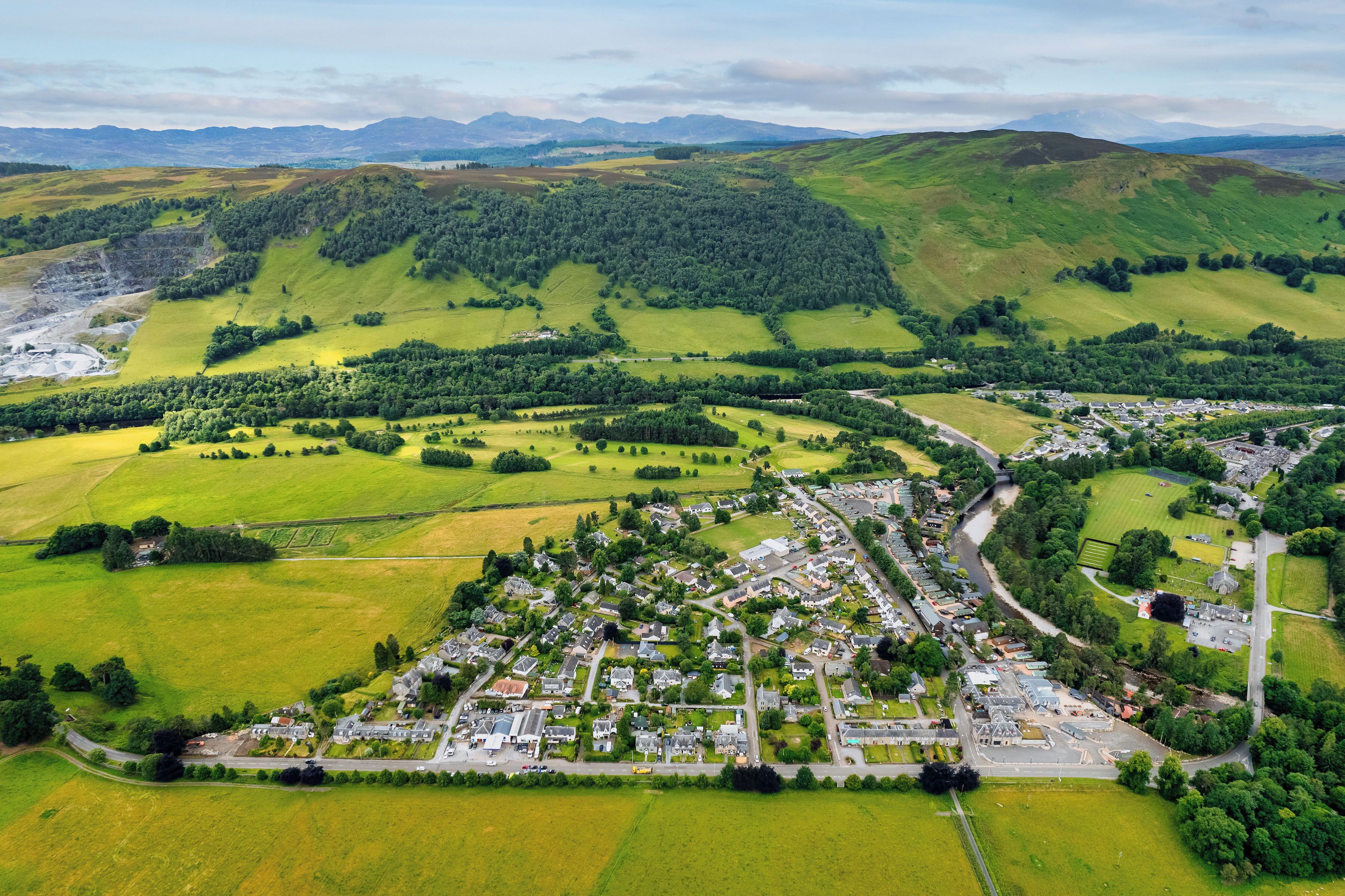 An aerial view of a town with a mix of residential and commercial buildings, surrounded by green fields and hills.