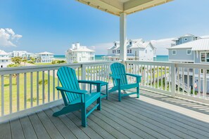 Une terrasse en bois avec deux fauteuils Adirondack turquoise, donnant sur une plage bordée de maisons blanches et sous un ciel d’un bleu limpide.