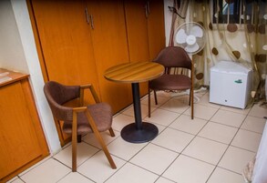 Una habitación pequeña con un armario de madera, una mesa redonda con dos sillas, un ventilador blanco y un refrigerador pequeño.