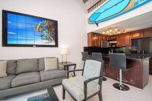Un espace de vie comprenant un canapé, un tabouret de bar, une table basse et un grand tableau encadré représentant une scène de plage.