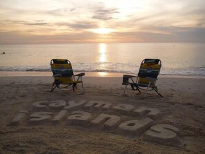 Two beach chairs facing the ocean with the text "Cayman Islands" written on the sand. - Beach Living at Island Pine Villas BLAW (Seven Mile Beach)