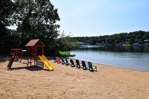 A sandy beach with a playground including a slide and climbing structure, and a row of colorful Adirondack chairs facing a calm lake with houses in the background.