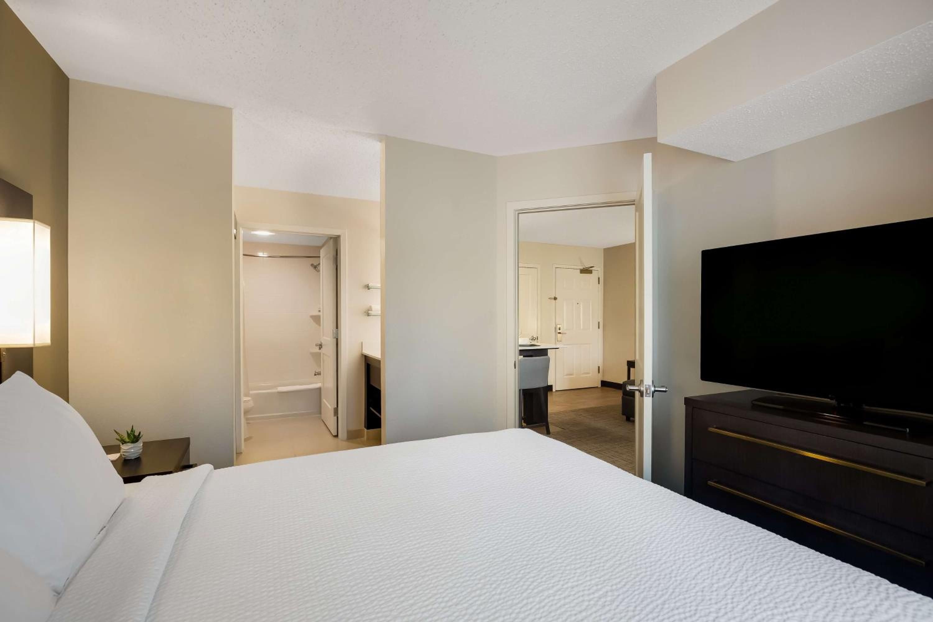 A hotel room with a large bed, a TV, a desk with a lamp, and a bathroom visible through an open door.