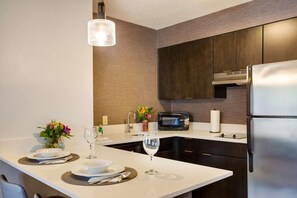 Een moderne keuken met donkerhouten kastjes, een roestvrijstalen koelkast, een magnetron, en een eethoek met een wit aanrecht en een bloevas.