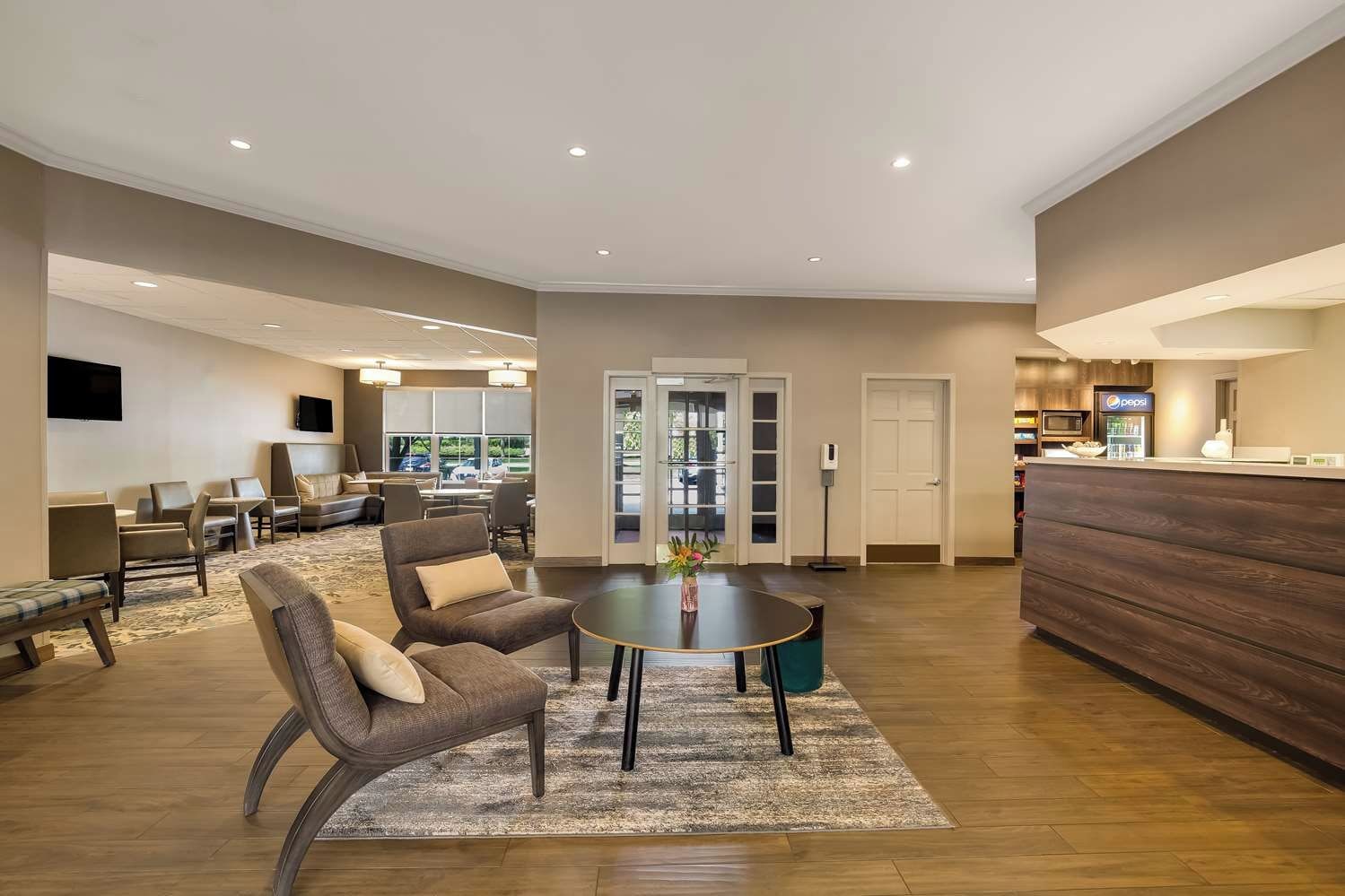 A spacious hotel lobby with a reception desk, seating area, and a view through the glass doors.