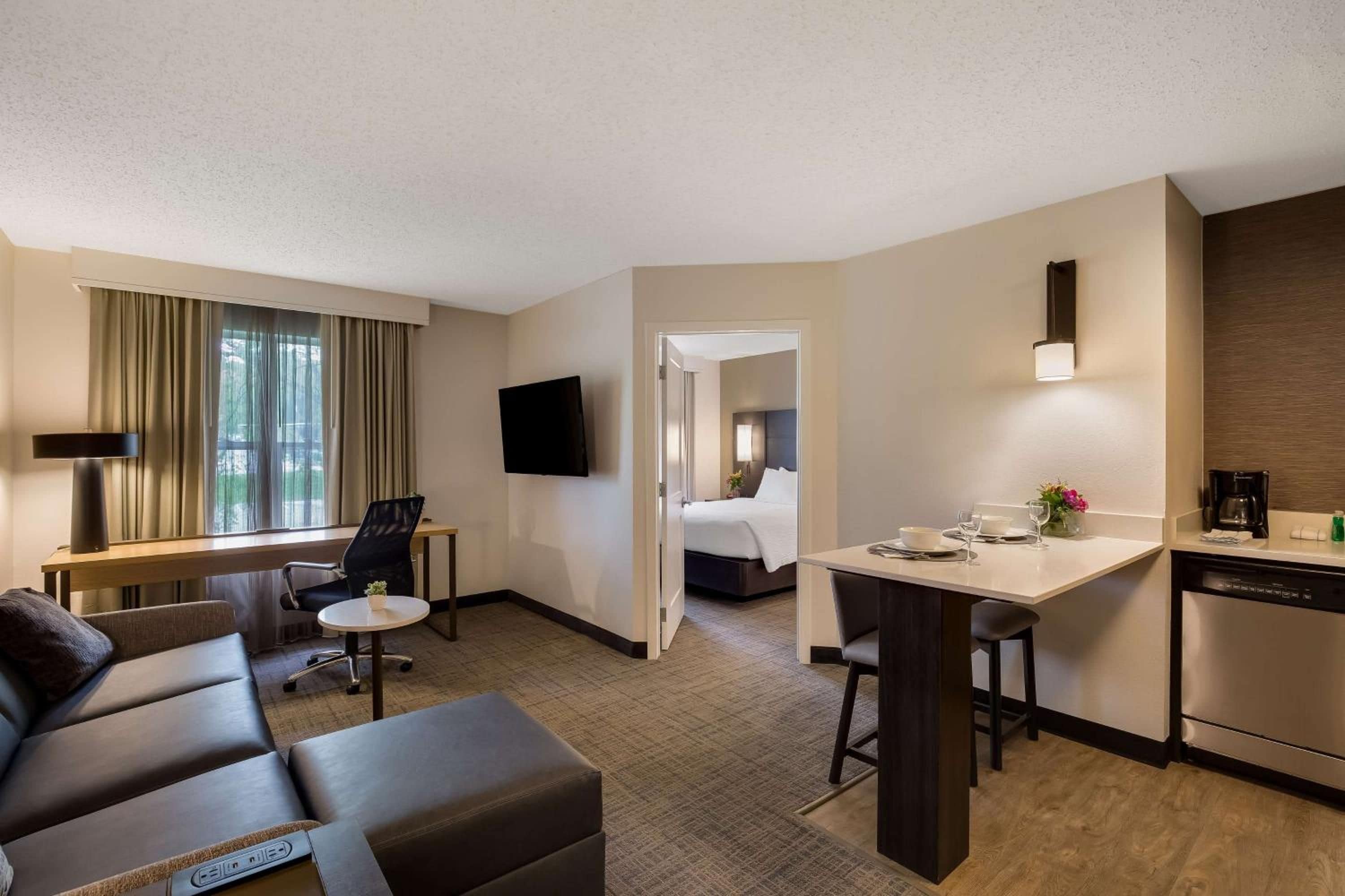 A hotel room with a sofa, a desk, a bed, a TV, and a kitchen area.