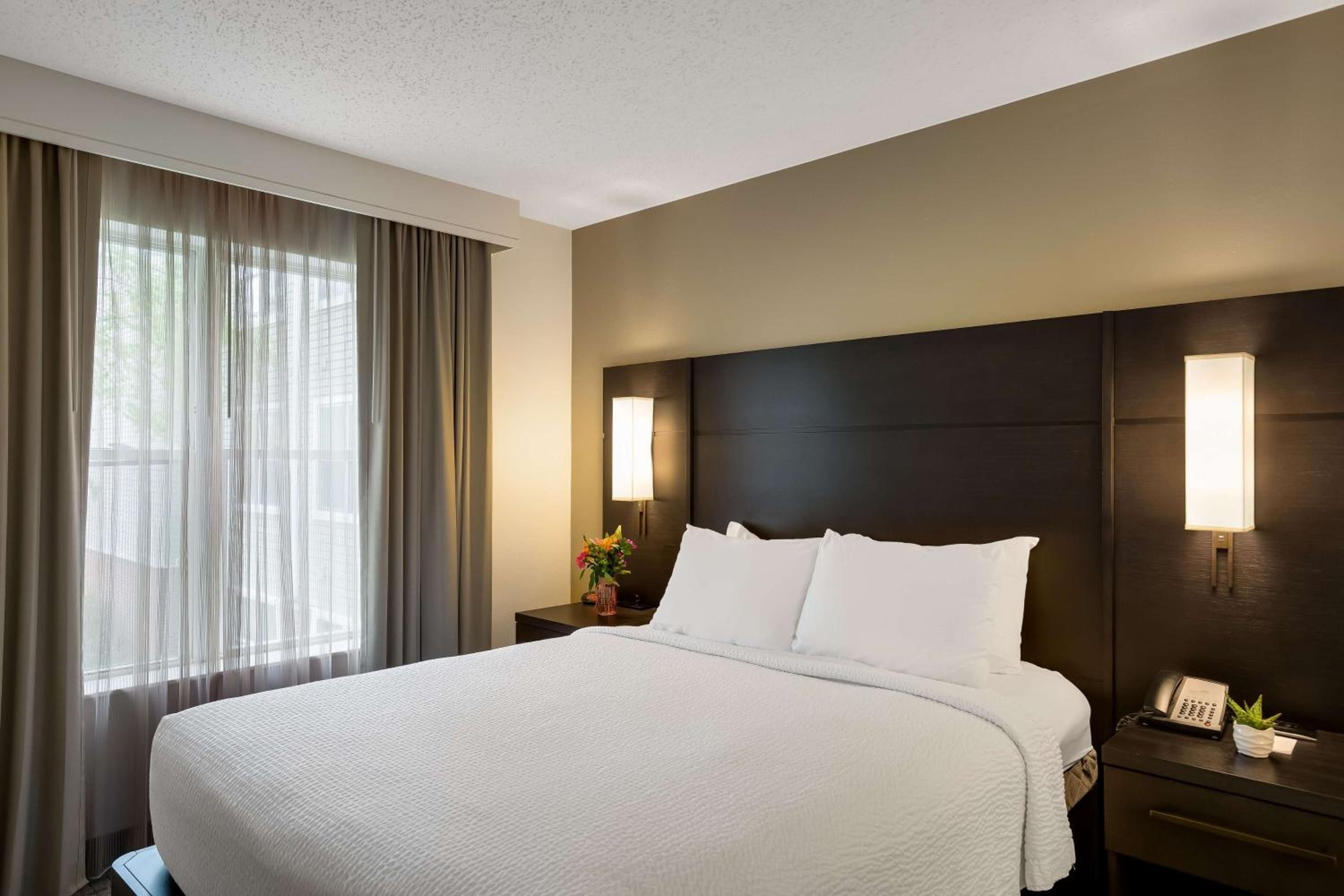 A hotel room with a large bed, two bedside tables, a nightstand, a lamp, and a window with curtains.