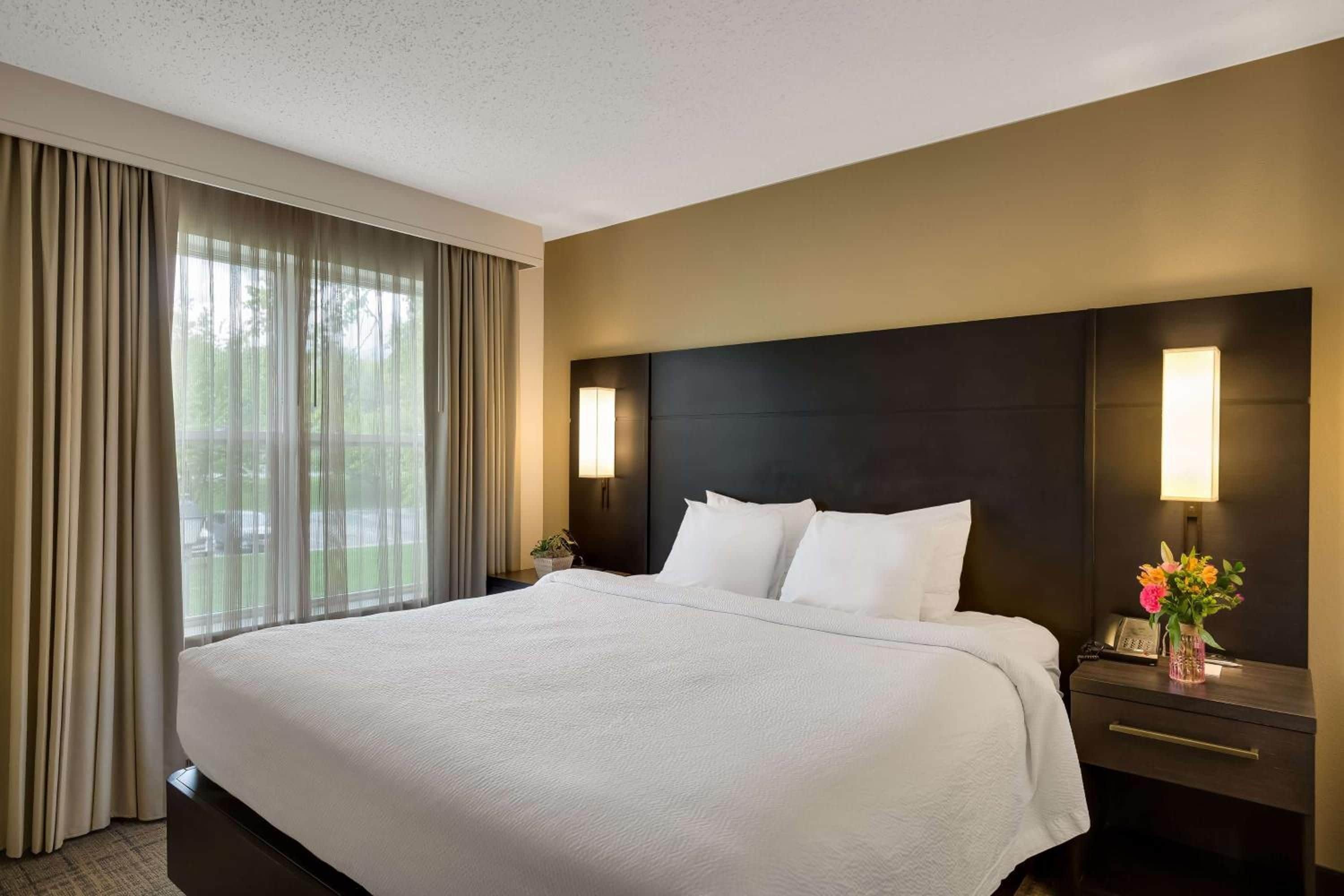 A hotel room with a large bed, a dark headboard, two bedside lamps, a nightstand with a phone, and a window with curtains.