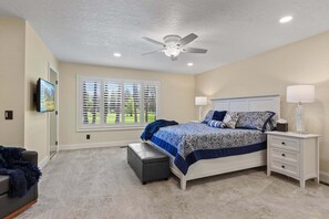 Une chambre à coucher comprenant un lit, un ventilateur de plafond, une table de chevet, une lampe, un banc, un téléviseur et une fenêtre avec des stores.