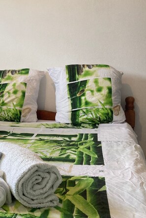 A bed with a green and white botanical print bedspread, two pillows with similar prints, and a rolled white towel. - Appart & Suites (Saint Laurent du Maroni)