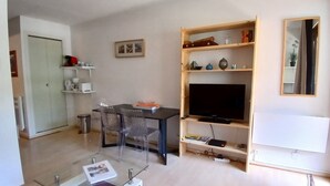 Una sala moderna con una mesa de centro de vidrio, un comedor con mesa y sillas, un televisor sobre un mueble de madera y una estantería con diversos objetos.