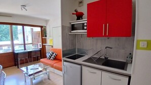 Cocina compacta con armarios blancos, un armario superior rojo y un microondas. Un tablero de centro de vidrio se encuentra en la sala, y un ventanal proporciona luz natural.