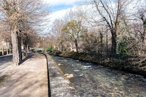 A clear river with a concrete walkway alongside it, surrounded by leafless trees and a stone wall.
