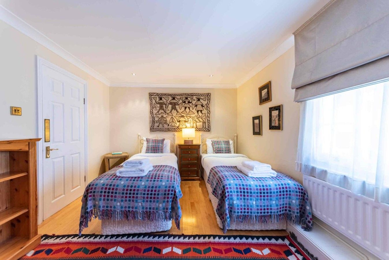 A room with two beds, a wooden floor, a patterned rug, a wall art piece, and a window with curtains.