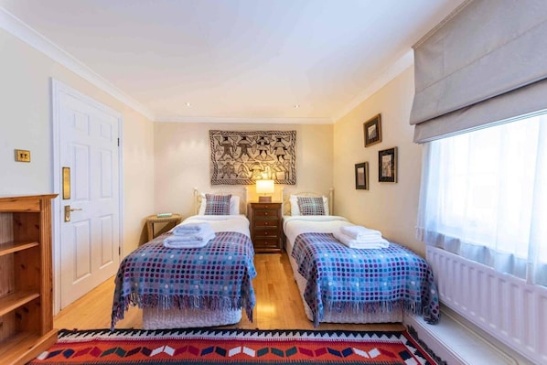 A room with two beds, a wooden floor, a patterned rug, a wall art piece, and a window with curtains. - Godfrey Street (London)