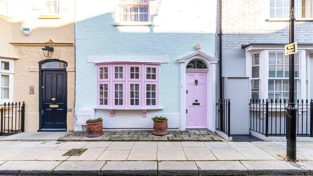 Um edifício com fachada azul clara, porta preta e janela rosa. Há duas plantas em vasos na frente.