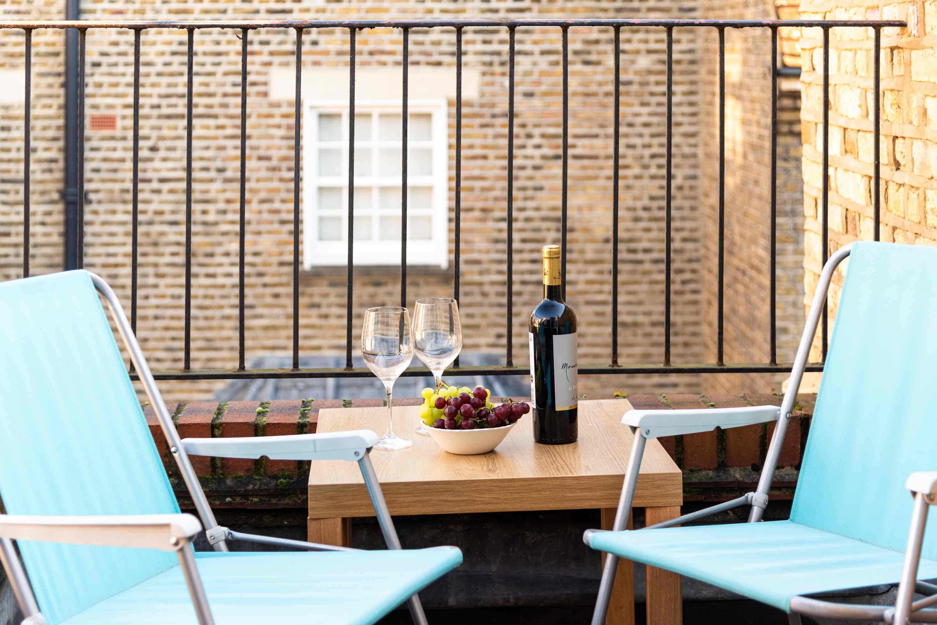 A table with a bottle of wine, two glasses, and a bowl of grapes on a balcony with brick walls and metal railings.