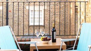 Uma mesa com uma garrafa de vinho, duas taças e uma tigela de uvas em uma varanda com paredes de tijolo e corrimão de metal.