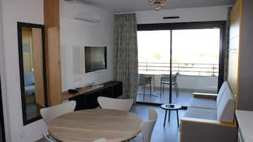 A modern living room with a round wooden table, white chairs, a flat-screen TV, and a sofa.