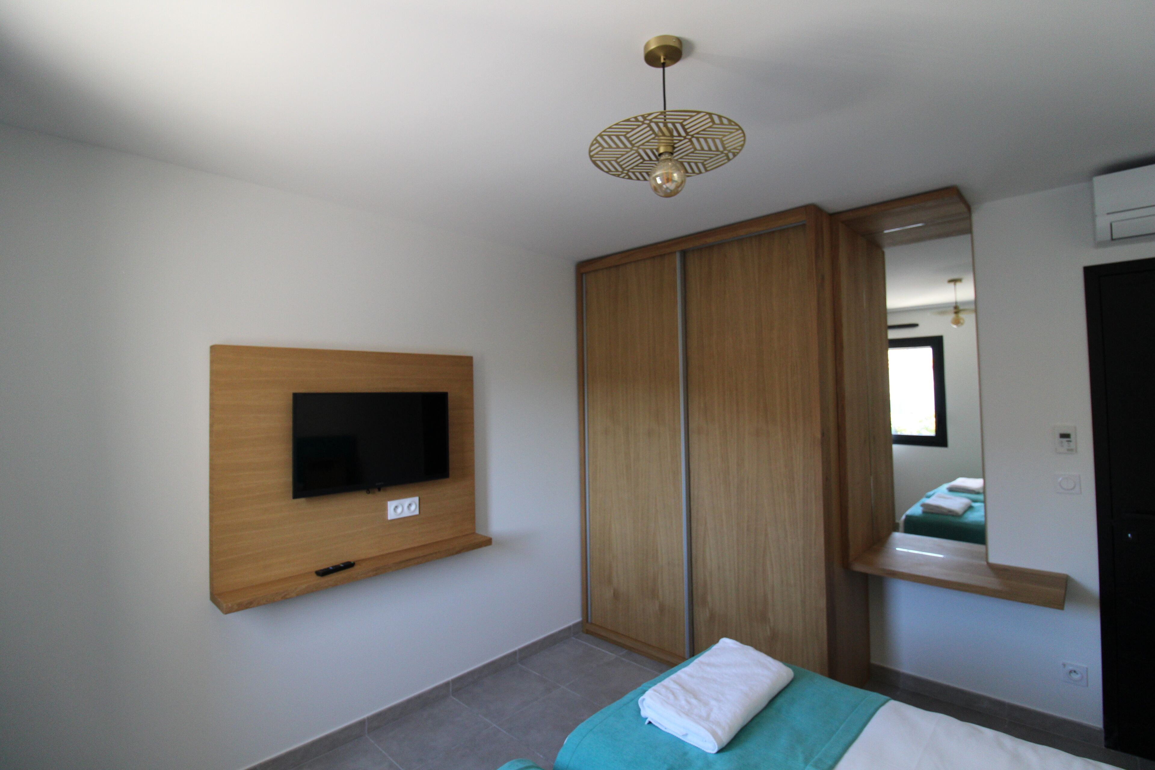 A bedroom with a TV mounted on the wall, a wardrobe, a bed with a green blanket, and a mirror above the bed.