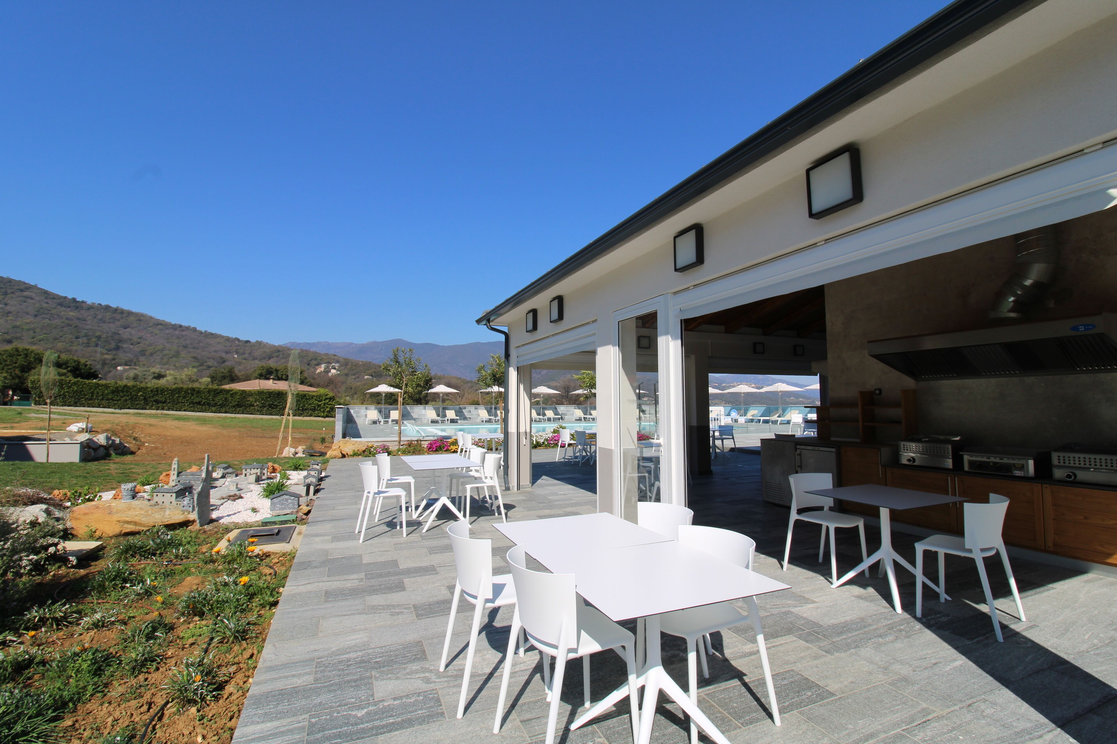 An outdoor patio with white tables and chairs, a pool area in the background, and a view of mountains.