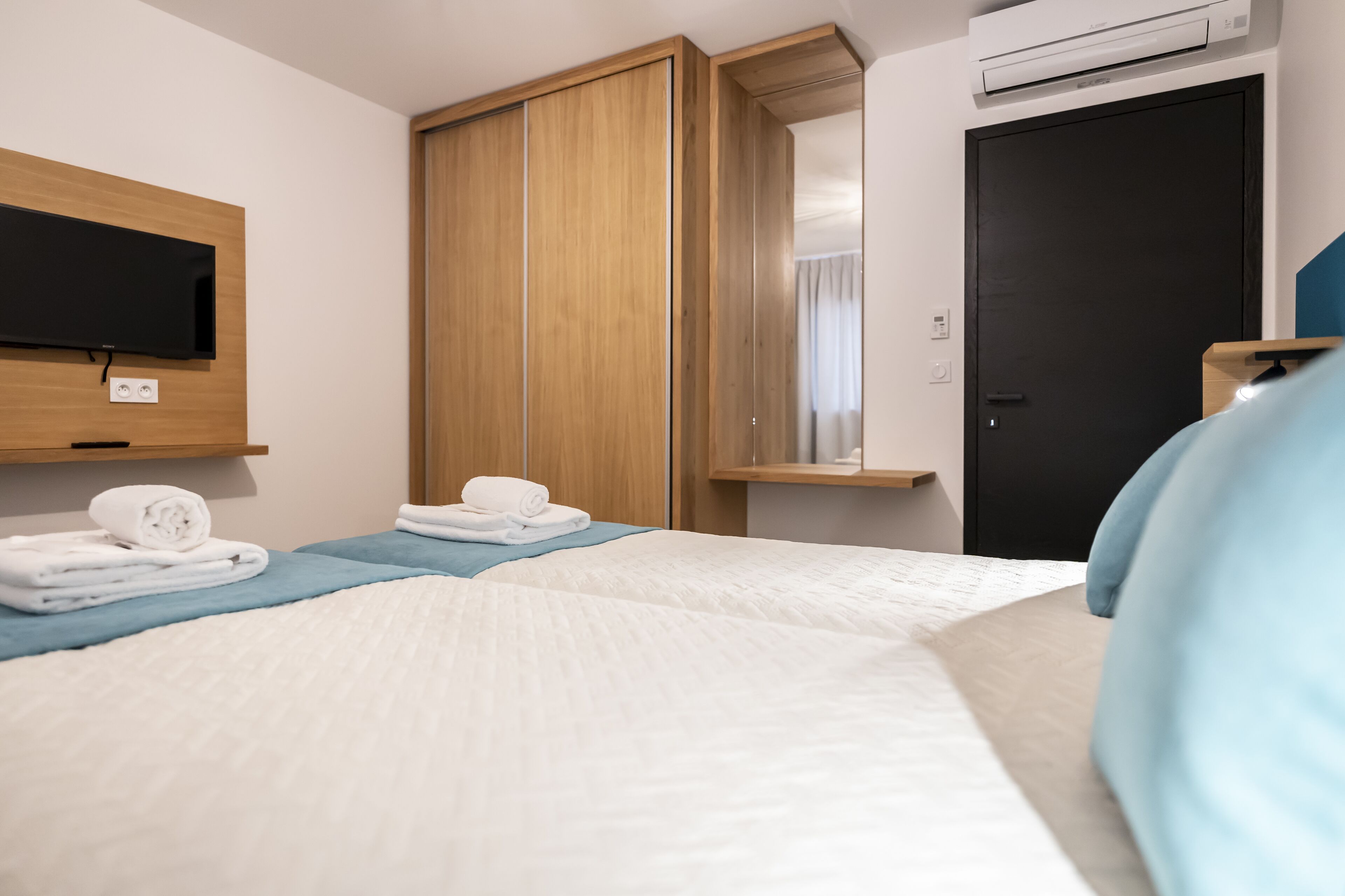 A hotel room with a bed, a TV mounted on the wall, a wardrobe, and an air conditioning unit.