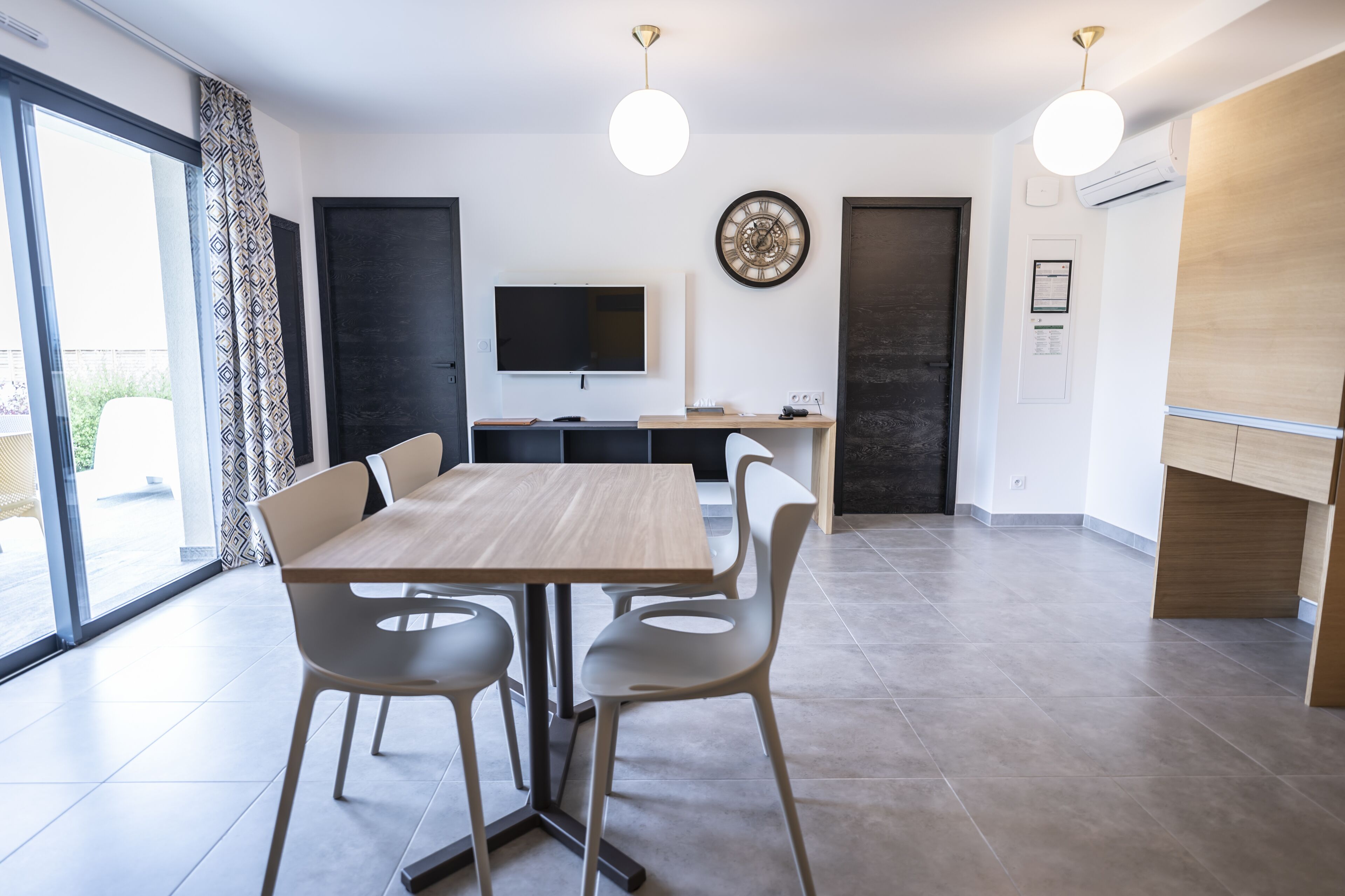 Un espace repas moderne avec une table et des chaises en bois, un téléviseur fixé au mur et un coin cuisine doté d’un plan de travail en bois.