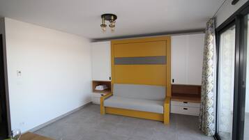Una habitación compacta con sofá, una mesita con vasos y una ventana con cortinas estampadas.