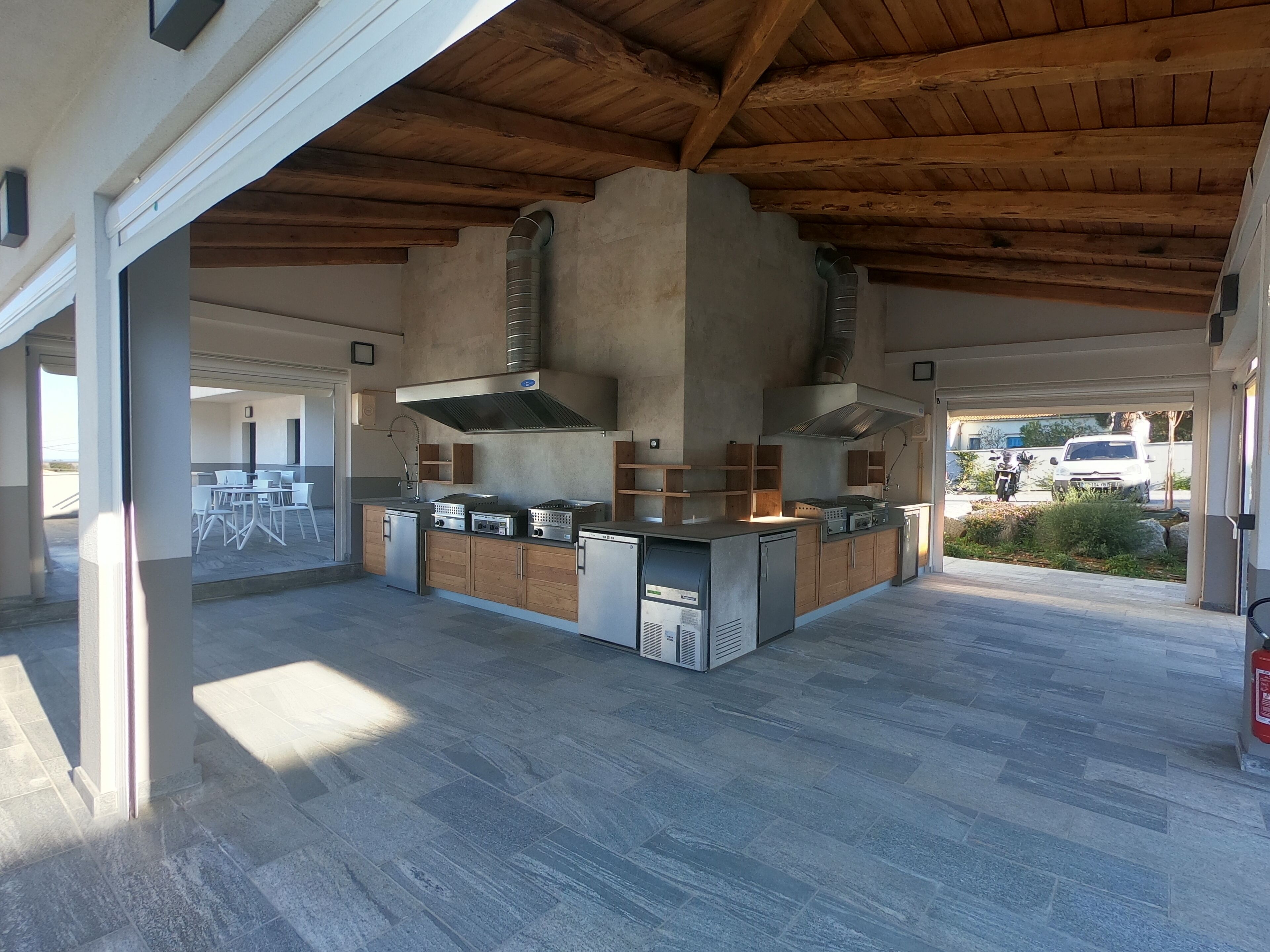 A spacious outdoor kitchen area with a high ceiling and exposed wooden beams, featuring multiple cooking stations and a seating area with tables and chairs.