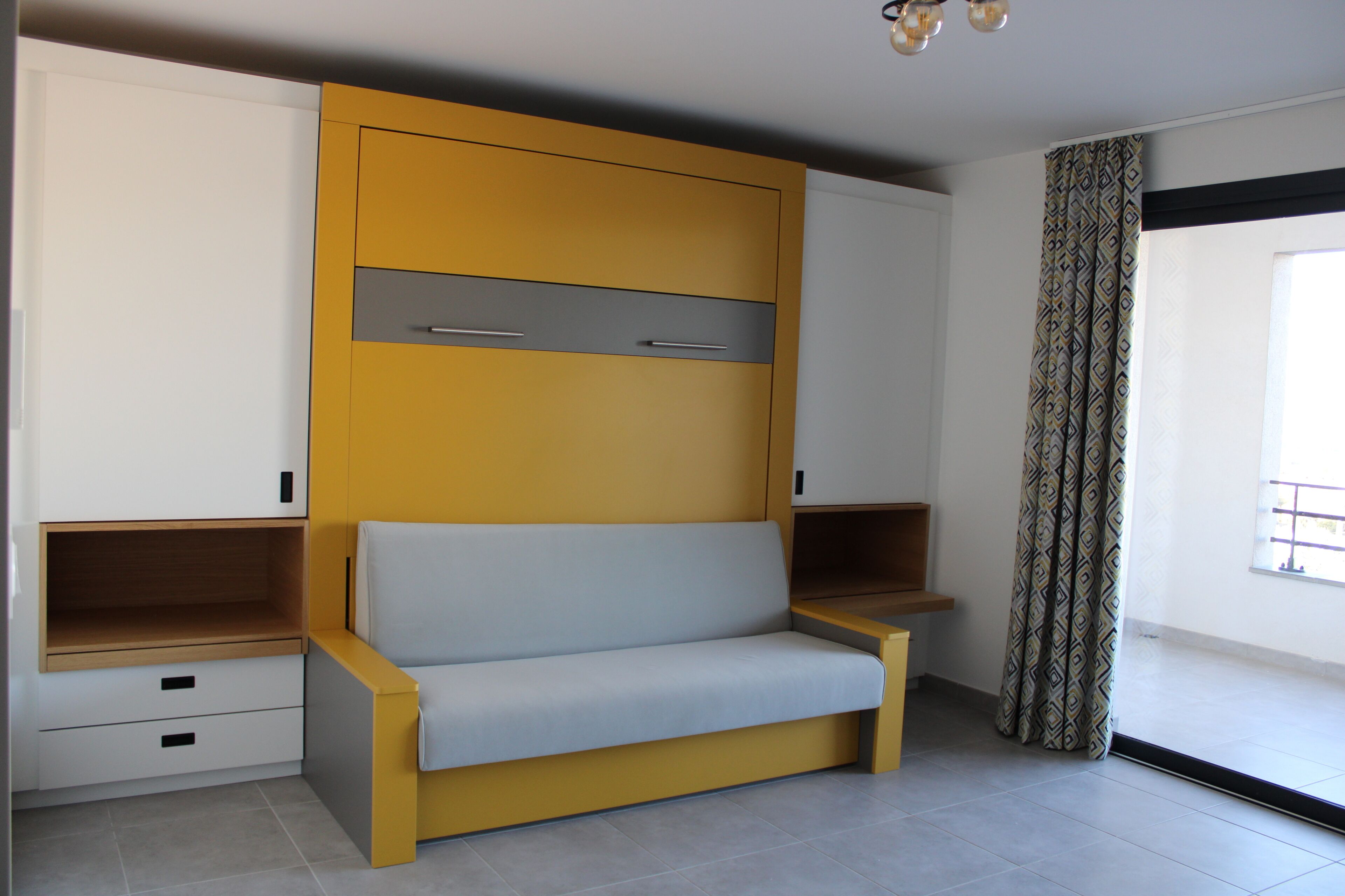 A modern room with a built-in sofa, storage cabinets, and a large window with patterned curtains.
