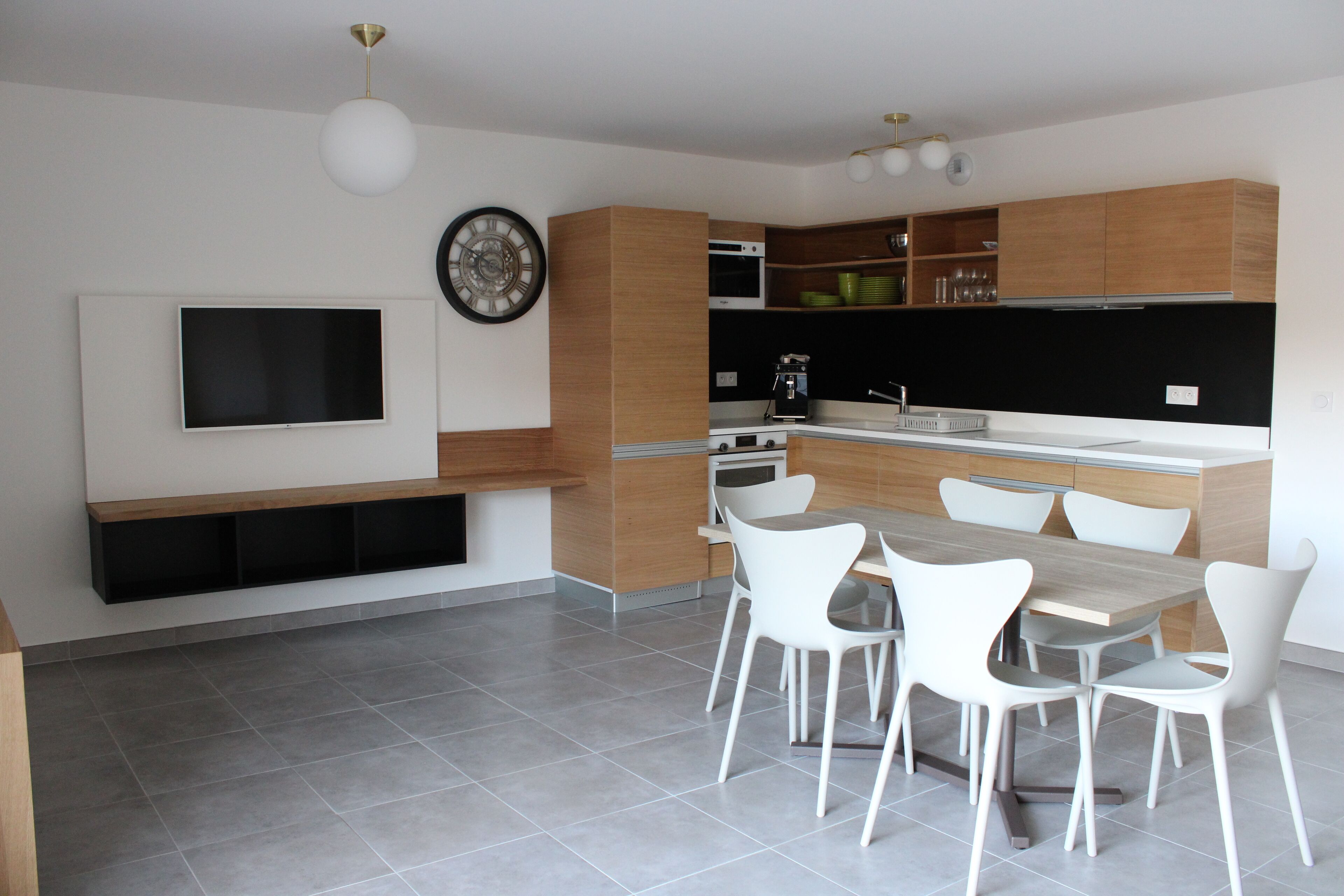 A modern kitchen with a built-in oven, microwave, and sink. There is a dining table with white chairs and a flat-screen TV mounted on the wall.