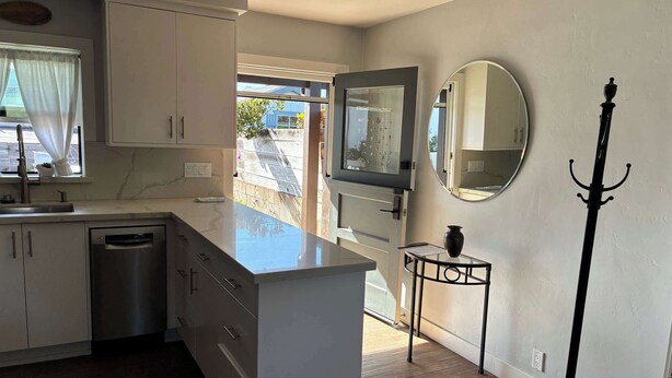 A modern kitchen with white cabinets, a built-in dishwasher, a large island, and a window with curtains. - Cozy Morro Bay Bungalo (Morro Bay)