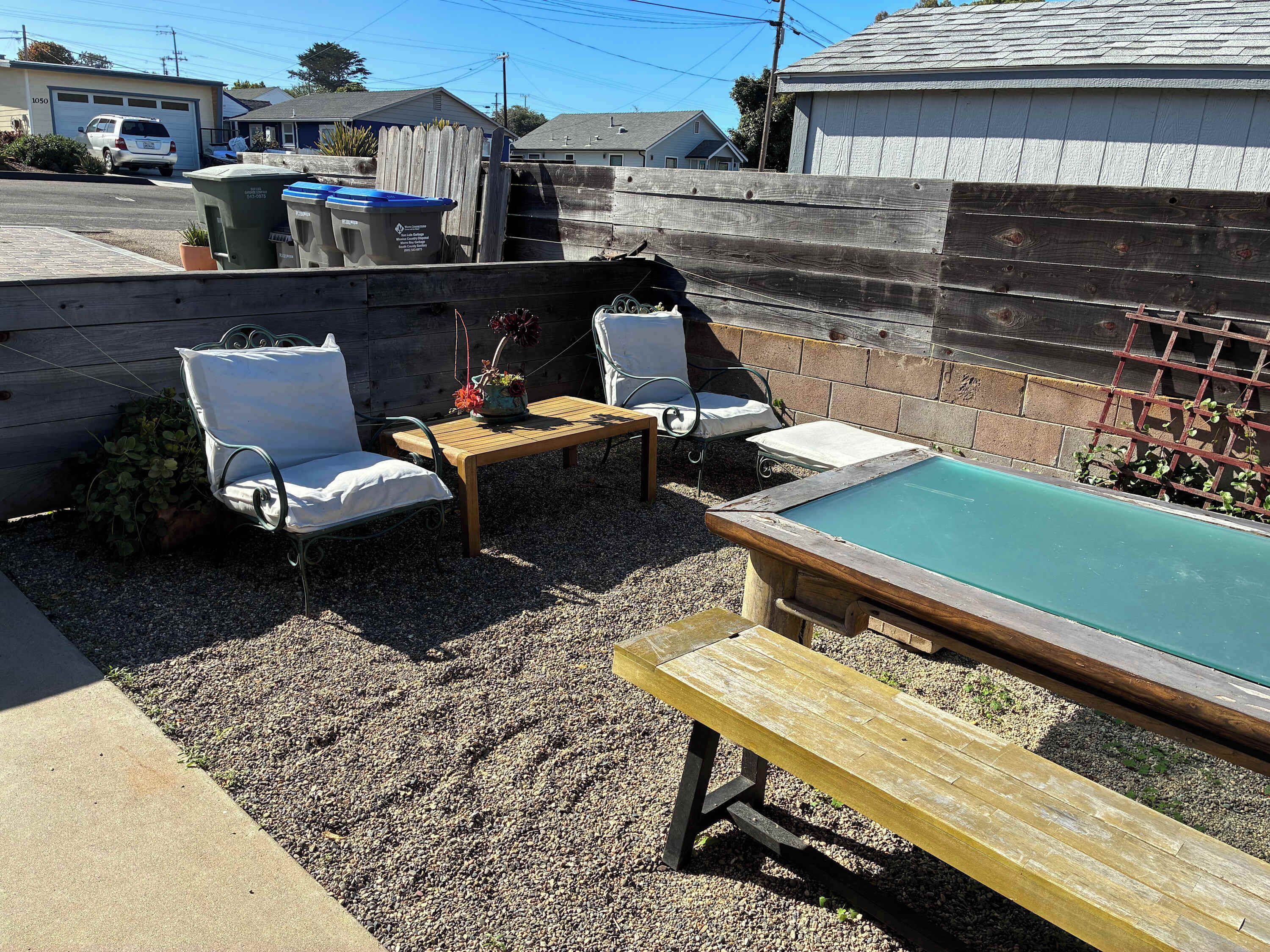 A backyard with a pool, two white lounge chairs, a wooden table, and a wooden bench.